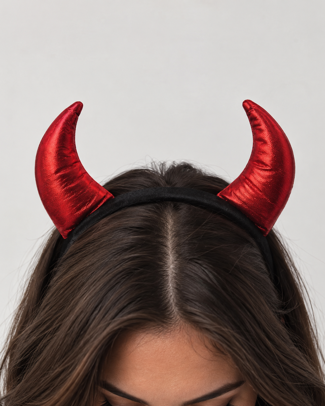 A close-up of a person wearing a black headband with two shiny red devil horns against a light gray background.