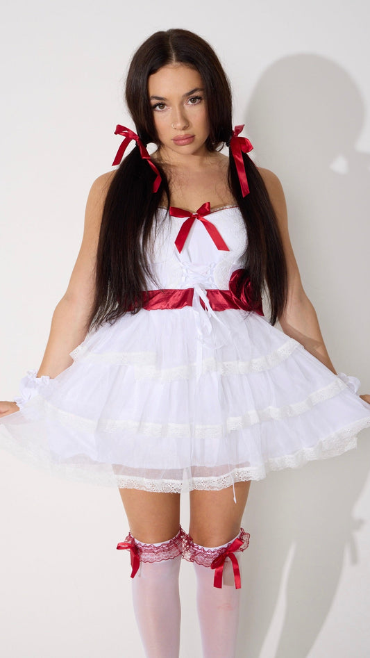 A woman modeling a white sleeveless tutu dress with red ribbon details, styled with white thigh-high stockings and red bows, against a plain white wall.